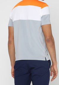 T-shirt à manches courtes en gris, blanc et orange. Présente un design en blocs de couleurs avec un col rond et des fentes latérales à l'ourlet.