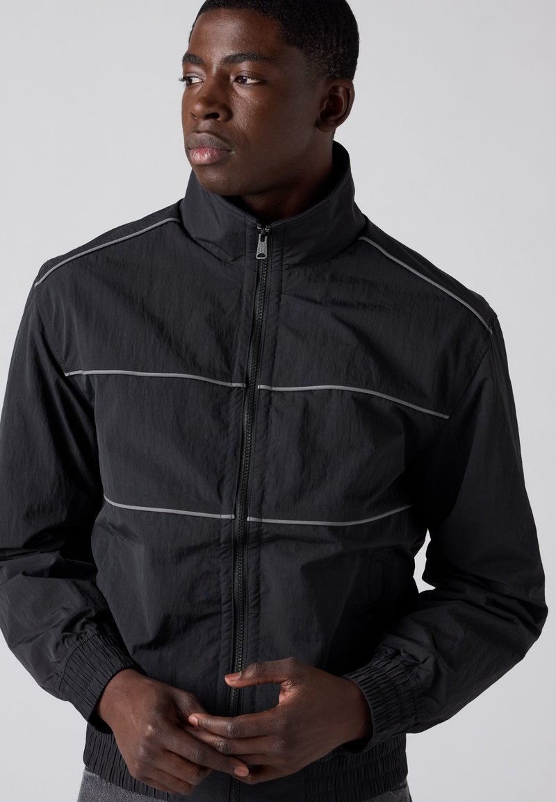 Jonge man die een zwart jack met ritssluiting en witte horizontale strepen draagt, kijkt naar links tegen een effen lichte achtergrond.