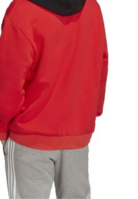 Felpa rossa con cappuccio nero, tessuto texturizzato e orlo a coste. Indossata con pantaloni della tuta grigi con strisce bianche laterali.