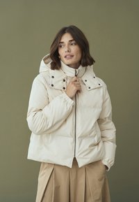 Purios spalvos pūkuota striukė su aukštu apykakle, spaudimo mygtukais ir elastinėmis rankovėmis. Striukė turi priekinį užtrauktuką ir gobtuvą.