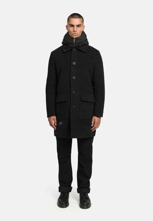 Black wool coat with a hood, featuring a zip closure and button-up front. It has two patch pockets and a slightly tailored fit. Black trousers and boots.