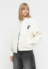 Veste bomber en polaire blanche avec des boutons-pression, des poignets côtelés et un logo brodé sur la poitrine gauche. Associée à un jean gris.