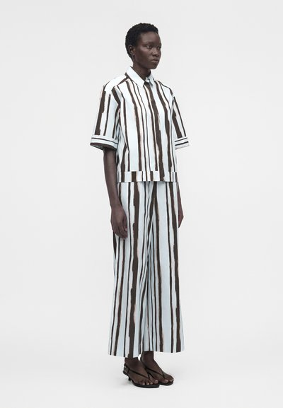 Άτομο που φορά χαλαρό πουκάμισο με κοντά μανίκια και γιακά και παντελόνια με φαρδιά πόδια με κατακόρυφες μαύρες και λευκές ρίγες, στέκεται μπροστά σε ένα απλό φόντο.