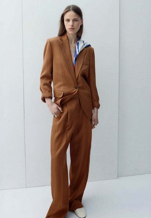 Grande femme aux longs cheveux bruns portant un costume marron oversize avec un pantalon à jambes larges et des chaussures blanches, debout devant un fond blanc uni.