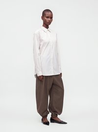 Personne debout portant une chemise blanche à fines rayures boutonnée, un pantalon marron ample avec des poignets élastiques, et des ballerines noires pointues sur fond blanc.