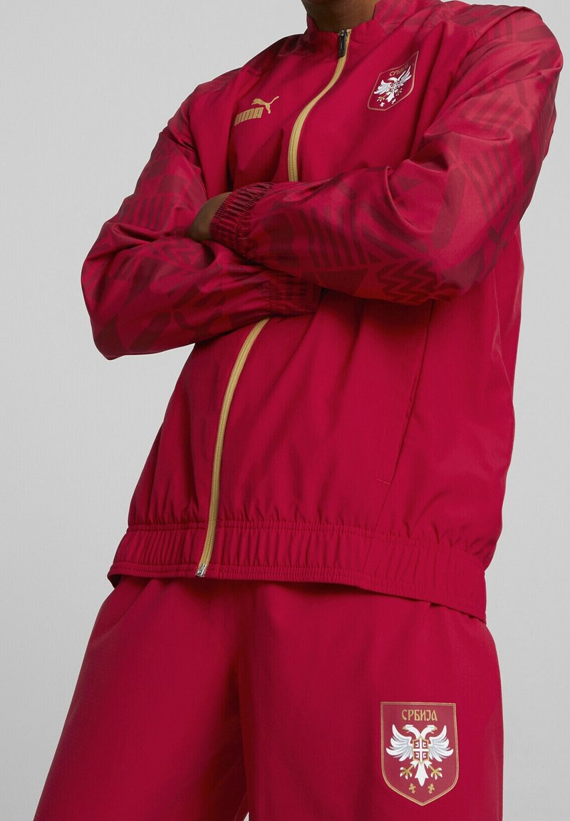 Survêtement de sport rouge en tissu léger, avec une fermeture éclair sur le devant, des poignets élastiques et un écusson représentant un aigle avec un texte en cyrillique sur le pantalon.