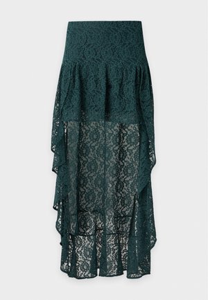 Un mannequin se tient devant un fond de studio uni, vêtu d’un haut noir, d’une jupe en dentelle vert foncé avec un long arrière et de chaussures plates noires.