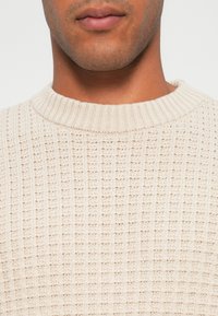 Maglione beige lavorato a maglia con un motivo testurizzato, dotato di un collo rotondo a coste. Il tessuto appare morbido e leggero.