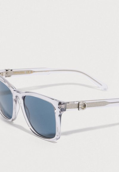 Διαφανή γυαλιά ηλίου με σκούρους μπλε φακούς, γεωμετρικό σχέδιο σκελετού, ασημί λεπτομέρειες στα μπράτσα και υφή στη μεντεσέ.
