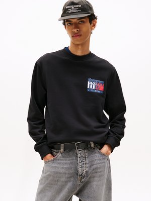 Felpa navy con logo frontale rosso e blu, abbinata a jeans denim grigi chiari. Il modello indossa un cappellino nero con scritta bianca e una cintura nera.