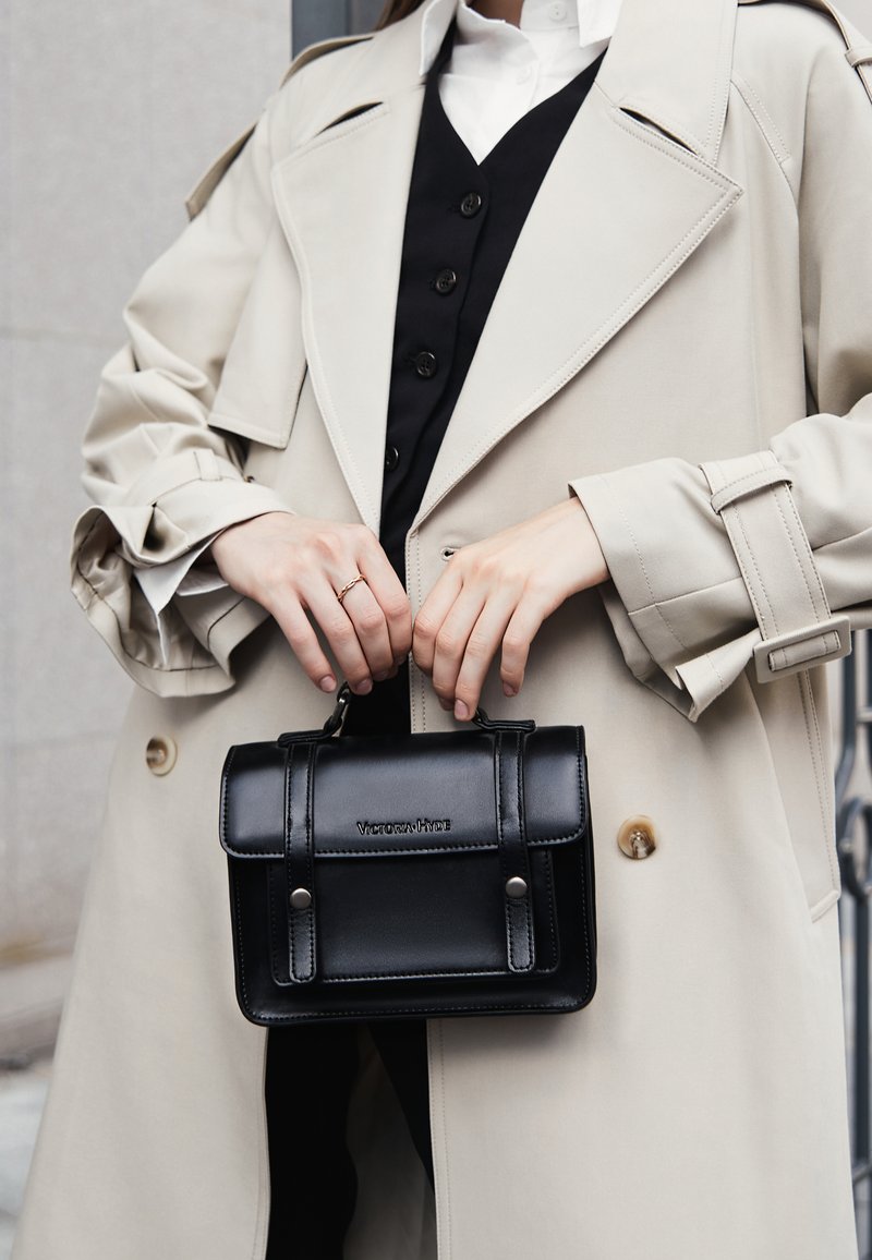 Borsa nera in pelle con un design strutturato, manico superiore e chiusura a scatto, indossata con un trench beige sopra un gilet nero.