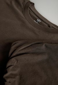 Divi tum�ši brūni T-krekli, kas izgatavoti no mīksta kokvilnas auduma, ar apaļu kakla izgriezumu un īsām piedurknēm, izklāti uz plaknes ar nedaudz redzamu tekstūras detaļu.