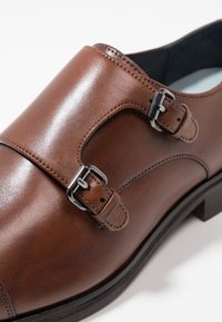 Zapato de vestir de cuero marrón con un acabado suave, que presenta dos hebillas plateadas y un diseño de punta redondeada, sobre una suela negra.