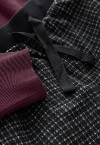 Tissu à carreaux noir et gris avec une manche côtelée bordeaux. Comprend des cordons de serrage noirs et une finition texturée.