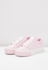 Różowe tenisówki z materiału typu canvas z białą gumową podeszwą, zaokrąglonym noskiem, sznurowadłami i metalowymi oczkami. Gładka faktura z delikatnym prążkowanym detalem na podeszwie.
