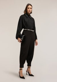 Blusa negra con cuello tachonado, pantalones negros ajustados, cinturón plateado y bolso negro; combinado con tacones de aguja negros.