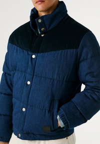 Modra podložena jakna z yoko iz temno modrega korduroja, z gumbi na pritiskanje, stranskimi žepi in rebrasto pletenimi manšetami. Ima teksturiran oblikovni vzorec.
