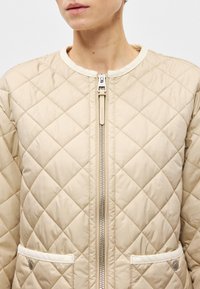 Μπεζ καπιτονέ μπουφάν με φερμουάρ μπροστά, ριμπ λαιμόκοψη και δύο μπροστινές τσέπες με φωτεινές λεπτομέρειες. Λείος υφασμάτινος σχε�διασμός με διαμαντένιο μοτίβο.