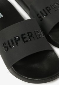 Infradito neri con una fascia texturizzata che presenta un logo "SUPERDRY" lucido nero. La suola è liscia e leggermente curvata.