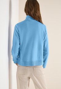 Femme aux longs cheveux bruns portant un haut à manches longues bleu clair et un pantalon beige, debout face à un fond clair uni.