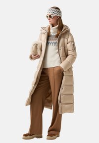 Beige pufferjack, chunky crème trui met zigzagpatroon, bruine wijde broek, bruine schoenen, witte zonnebril en haarband.