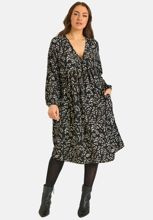 Kobieta ubrana w czarną sukienkę do kolan z beżowymi abstrakcyjnymi plamami, z długimi rękawami i dekoltem w serek, czarne rajstopy i czarne botki, stoi z jedną ręką w kieszeni.