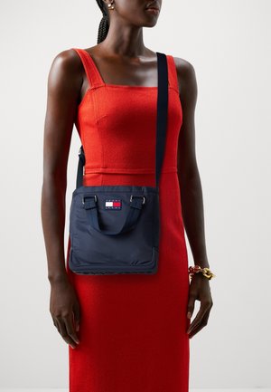 Granatowa nylonowa torba na ramię z podwójnymi uchwytami i regulowanym paskiem, o prostokątnym kształcie i naszywce z logo. Gładka faktura.