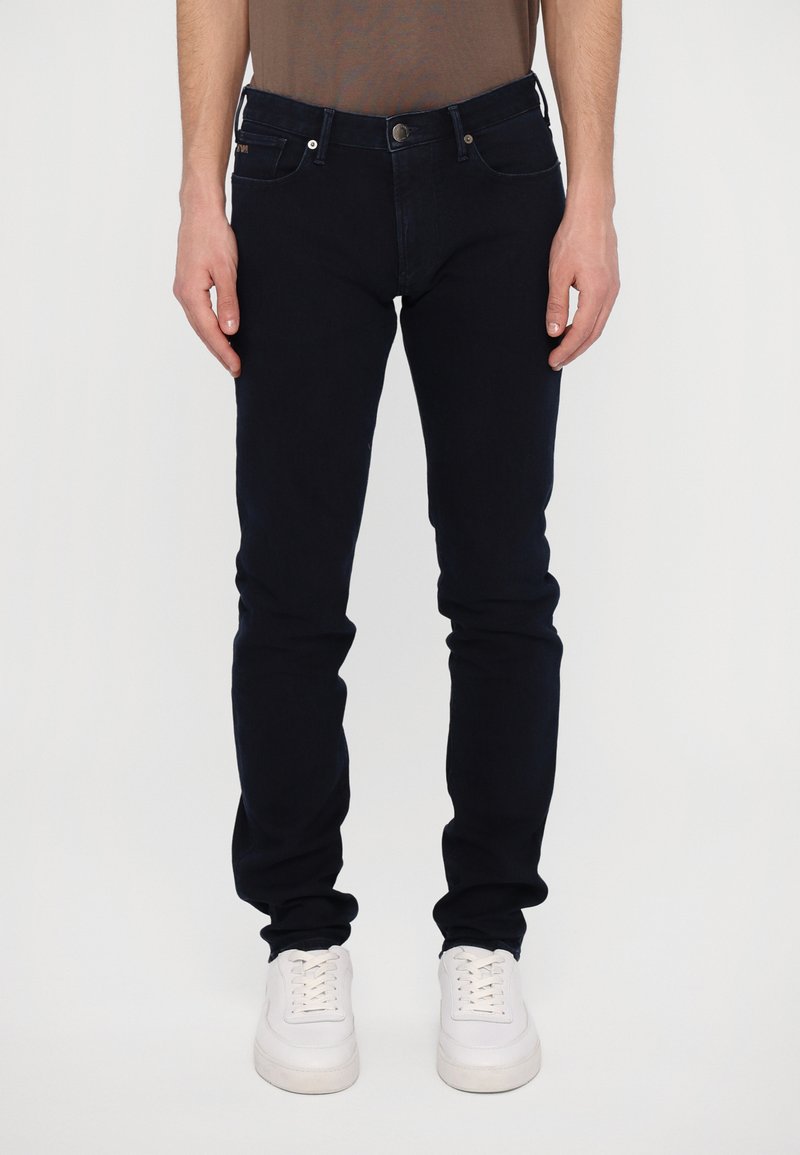 Dark blue skinny jeans made of denim, featuring a standard button and zipper closure, five pockets, and a subtle stitched logo on the back pocket.