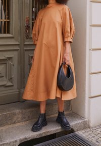 Hnědé bavlněné šaty s volným střihem, balónovými rukávy a knoflíkovými detaily. Spárováno s černými kotníkovými botami a černou zakřivenou kabelkou.