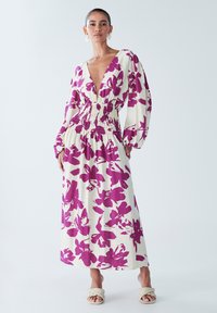 Robe maxi en crème avec un grand motif floral violet, décolleté en V profond, taille élastique et manches bouffantes longues. Portée avec des sandales tressées.