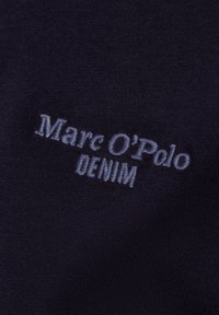 Donkere stof met een gladde textuur, waarop in het lichtblauw geborduurde tekst "Marc O'Polo DENIM" prominent is weergegeven.