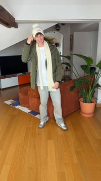 Grøn parka med pelskrave, hvid beanie med logo, cremefarvet tørklæde, hvid t-shirt, lyse jeans, sølvfarvede sneakers, varm indendørs atmosfære, potteplante.