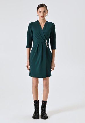 Robe portefeuille turquoise avec des manches 3/4, ornée d'une boucle et d'une chaîne à la taille, associée à des bottines à lacets noires. Finition en tissu lisse.