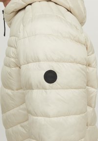 Light beige puffer jacket with quilted sections, featuring a black circular logo patch and a drawstring hood. Smooth, glossy texture.
