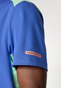 Blue and green short-sleeve sports shirt on arm with orange "ASICS PADEL" logo on the sleeve against a white background.