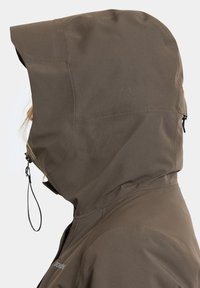 Widok z boku osoby w brązowej kurtce z kapturem z regulowanym ściągaczem na kapturze, na jednolitym tle.