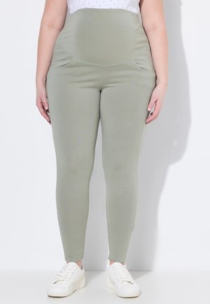 Kobieta ubrana w jasnozielone legginsy z wysokim stanem i bocznymi kieszeniami oraz białe trampki, stojąca na białym tle.