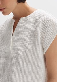 Witte top met een textuur, geweven patroon. Beschikt over een zachte V-hals en korte mouwen, wat een modern, minimalistisch ontwerp benadrukt.