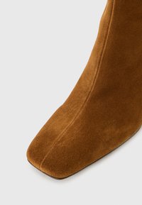 Bota de tobillo de ante marrón con diseño de punta cuadrada, textura suave y detalles de costura limpia. La suela parece plana sin hardware visible.