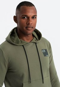 Felpa verde oliva in morbido misto cotone, caratterizzata da una tasca frontale, lacci neri con accenti bianchi e una patch con logo sul petto.
