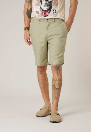 Man draagt een beige t-shirt met schedelprint, lichtgroene korte broek tot op de knie, beige instappers en toont beentattoeages tegen een neutrale achtergrond.