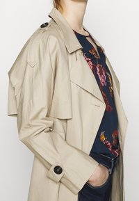 Cappotto beige con tasche a pattina, spalline e bottoni neri. Indossato sopra una maglia floreale blu navy. Tessuto testurizzato con cuciture visibili.