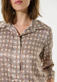 Seidenbluse mit geometrischem Muster in Beige-, Braun- und Blautönen, mit spitzem Kragen und Knopfdetails auf der Vorderseite.
