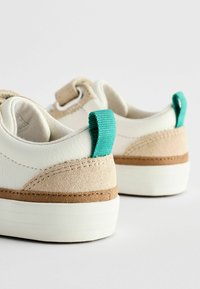 Weiße Lederschuhe mit beigen Wildlederakzenten und türkisfarbenen Stoffschlaufen, ausgestattet mit einer strukturierten Sohle und einer kontrastierenden braunen Zwischensohle.