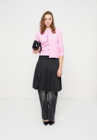 Ροζ πουλόβερ με κουμπιά, μαύρη πλισέ φούστα, γκρι παντελόνι με φαρδιά πόδια, μαύρες γόβες με μυτερές μύτες, κρατώντας μια μικρή μαύρη τσάντα.