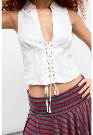 Haut court sans manches blanc avec col et détail lacé à l'avant, associé à une jupe taille haute grise avec des rayures horizontales rouges.
