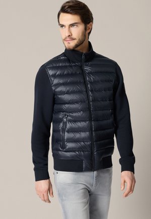 Mężczyzna w granatowej pikowanej kurtce puchowej z kieszeniami na suwak oraz jasnoszarych dżinsach, stojący na tle jednolitego beżowego tła.