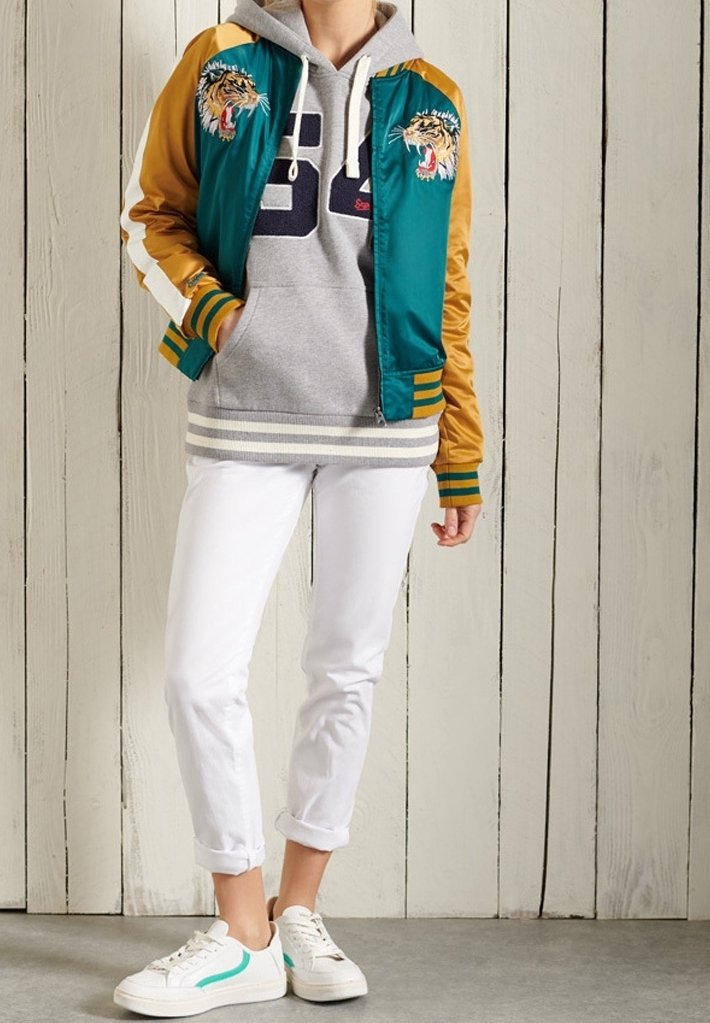 Personne portant une veste bomber bleu sarcelle et moutarde avec une broderie tigre, un sweat à capuche gris, un pantalon blanc retroussé et des baskets blanches devant un mur en bois.