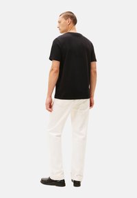 Camiseta de algodón negra con mangas cortas, escote redondo, combinada con pantalones blancos de pierna recta y zapatos negros, vista desde la parte de atrás.
