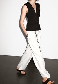 Svart ärmlös väst med figurnära design och knappar framtill, kombinerad med vida vita byxor och svarta sandaler, vilket skapar en kontrasterande look.